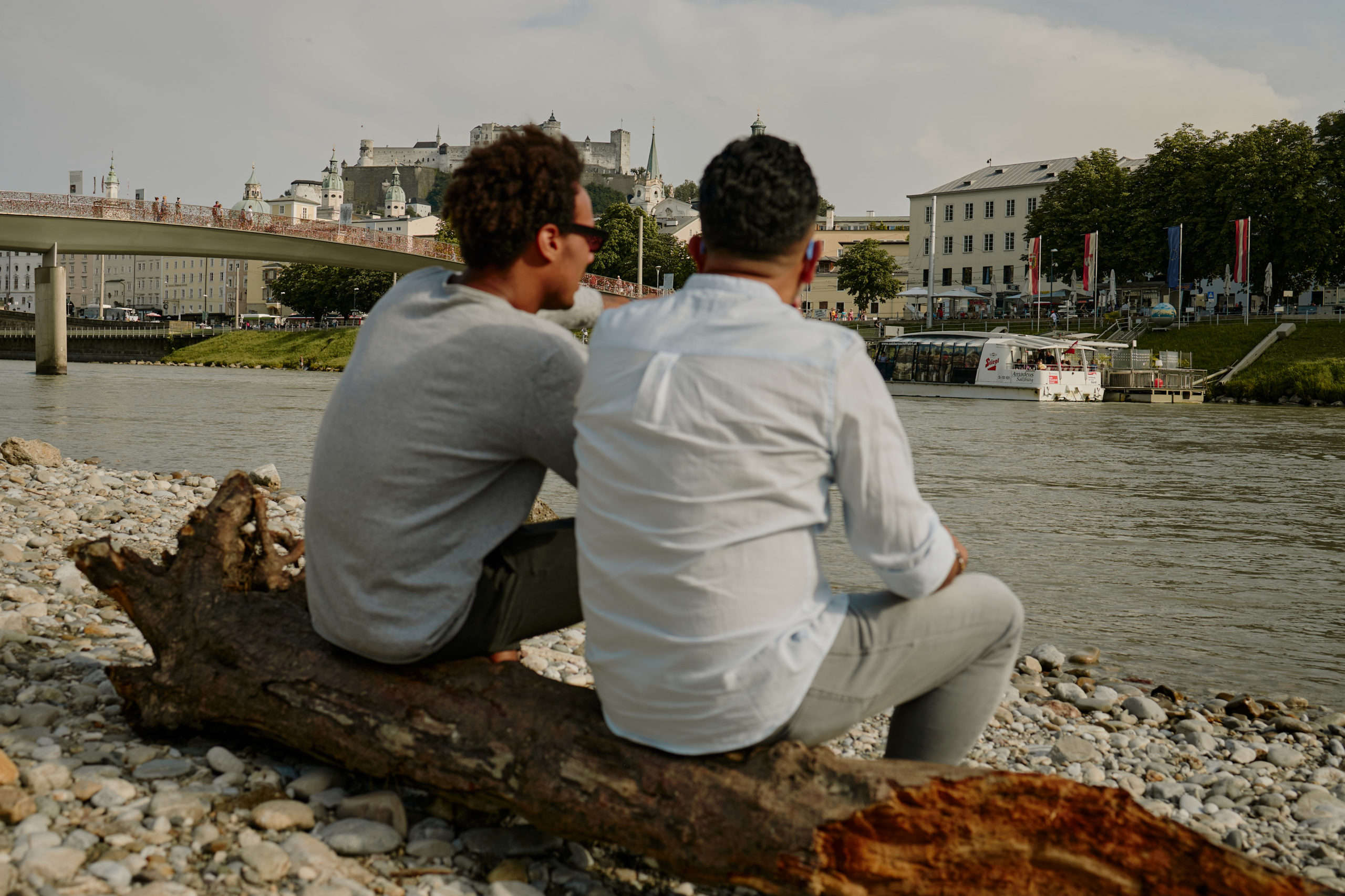 Zwei Männer sitzen auf einem Baumstamm und schauen auf die Salzach, den Fluss durch Salzburg